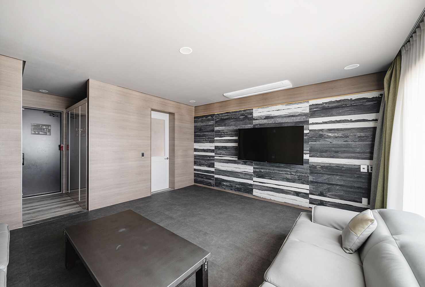 A modern hotel room with a flat-screen TV mounted on a textured wall, a sofa, a small table, and a door leading to a closet.