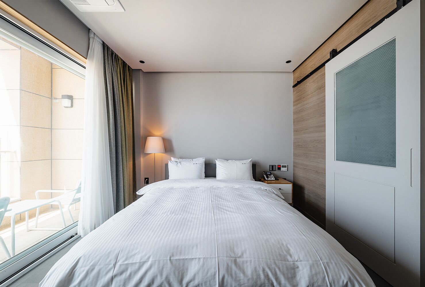 A hotel room with a large bed, a bedside table with a lamp, a phone, and a view of the outside balcony.
