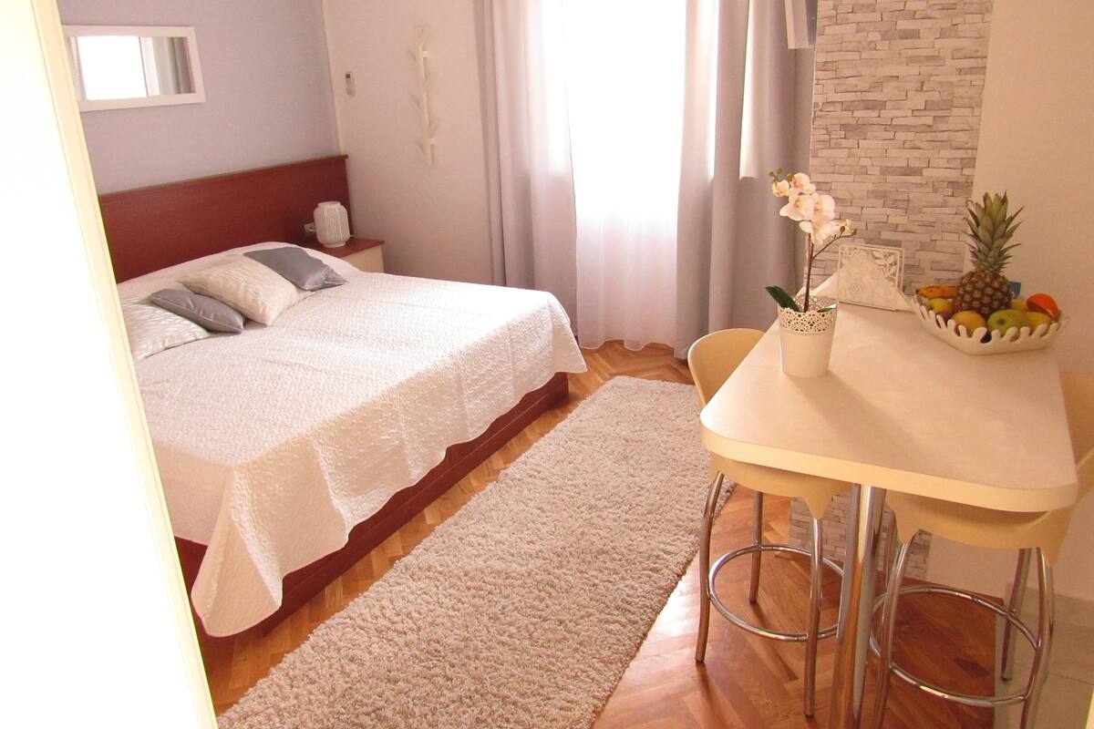 Une chambre à coucher avec un lit, une table de chevet, une table avec des fruits et une fenêtre avec des rideaux.