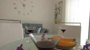 A dining area with a table set for two, featuring a martini glass and a plate of melon. A clock is visible on the wall, and a couch is in the background. - Apartmani Roko (Kastela)
