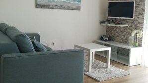 A modern living room with a grey sofa, a white coffee table, a flat-screen TV on a stone-patterned wall, and a painting of the ocean. - Apartmani Roko (Kastela)