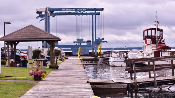 A marina with a wooden dock, a covered waiting area, and a large blue crane labeled "Washburn Marina" in the background. - Driftwood Cottage: Your Gateway to Lake Superior & Apostle Islands (Washburn)