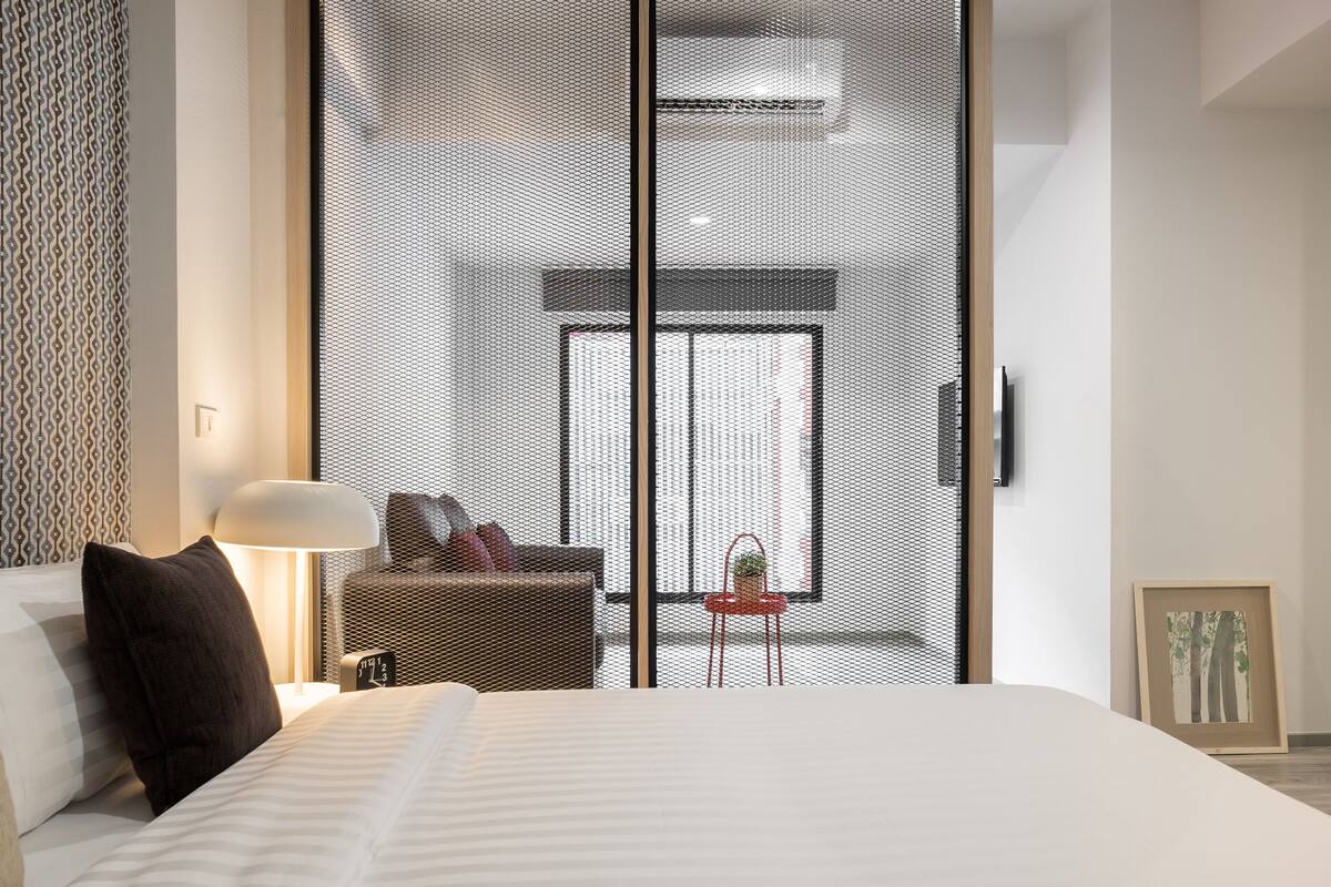 A modern hotel room with a large bed, a small side table with a lamp, a wicker chair, and a view through a large window.