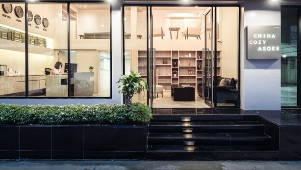 Un edificio moderno con entrada de vidrio, un letrero que dice "CHINA COZY ASOKE" y un interior visible con asientos y estanterías.