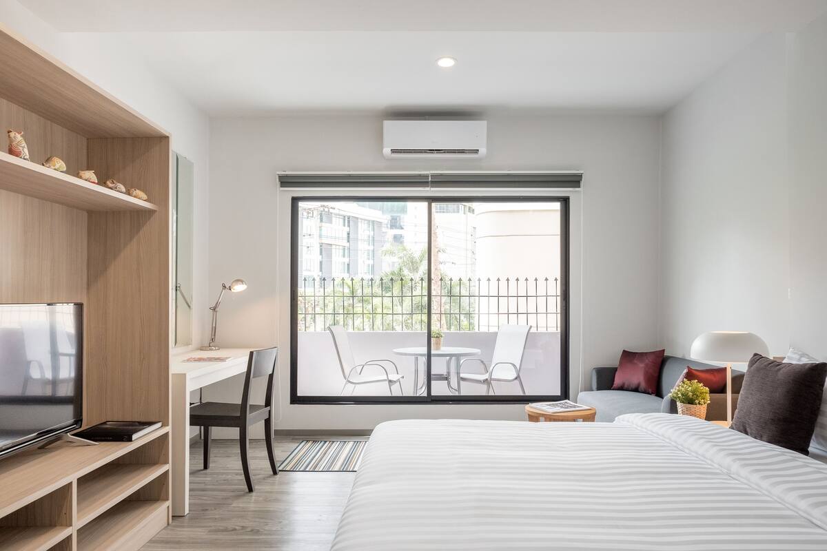 A modern hotel room with a large bed, a desk with a chair, a television, and a balcony with a table and chairs.