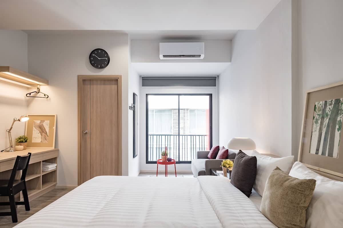 A modern hotel room with a large bed, a desk with a chair, a small table with a plant, a sofa with cushions, a clock, and a window with blinds.