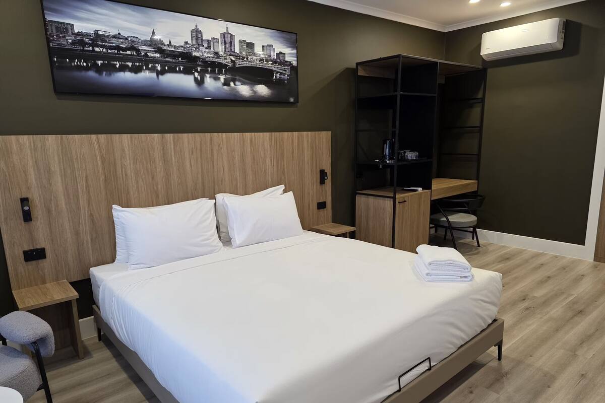 Une chambre d’hôtel avec un lit, un bureau, une chaise, une armoire et un tableau représentant un paysage urbain accroché au mur.