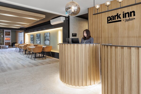 A reception area with a wooden reception desk, a receptionist, and a seating area with chairs. - Park Inn By Radisson Melbourne Carlton (Melbourne)