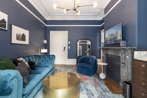 Uma sala de estar moderna com um sofá azul, uma mesa de centro redonda dourada e uma pintura de paisagem urbana na parede.