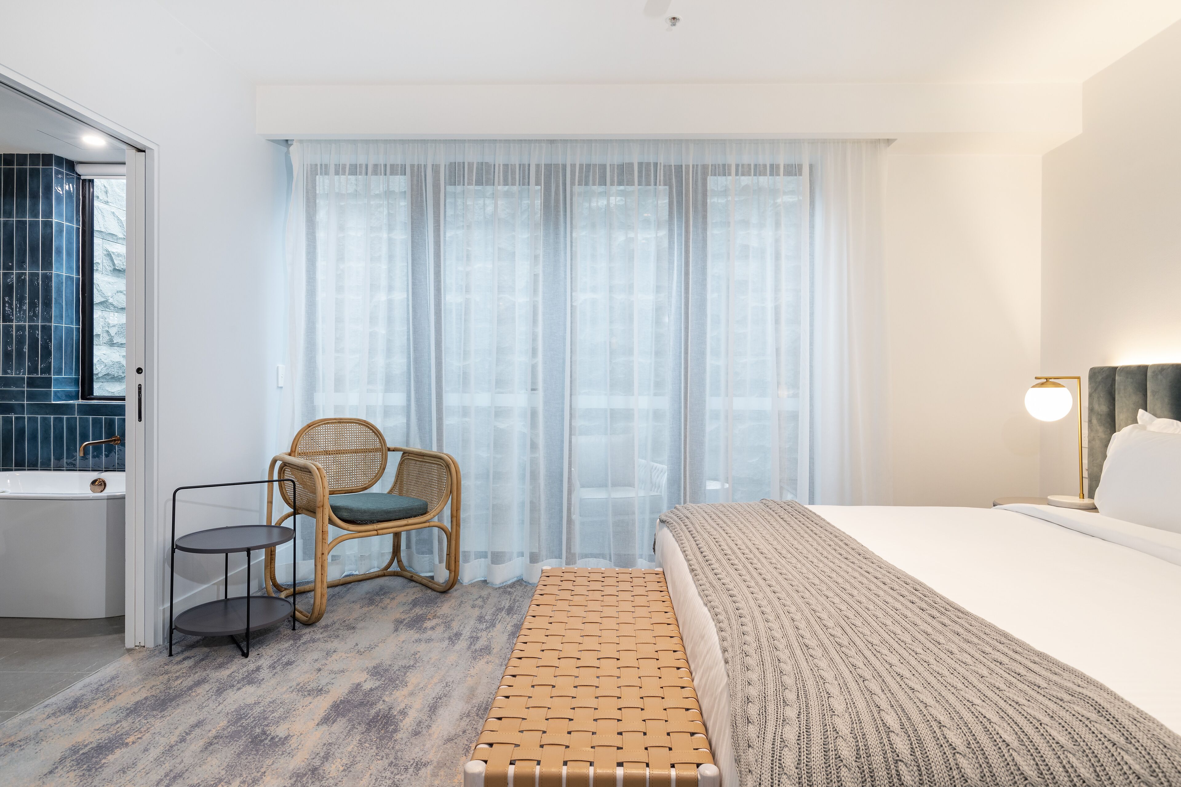 Una habitación de hotel con cama, una silla, una mesita y una ventana con cortinas traslúcidas.