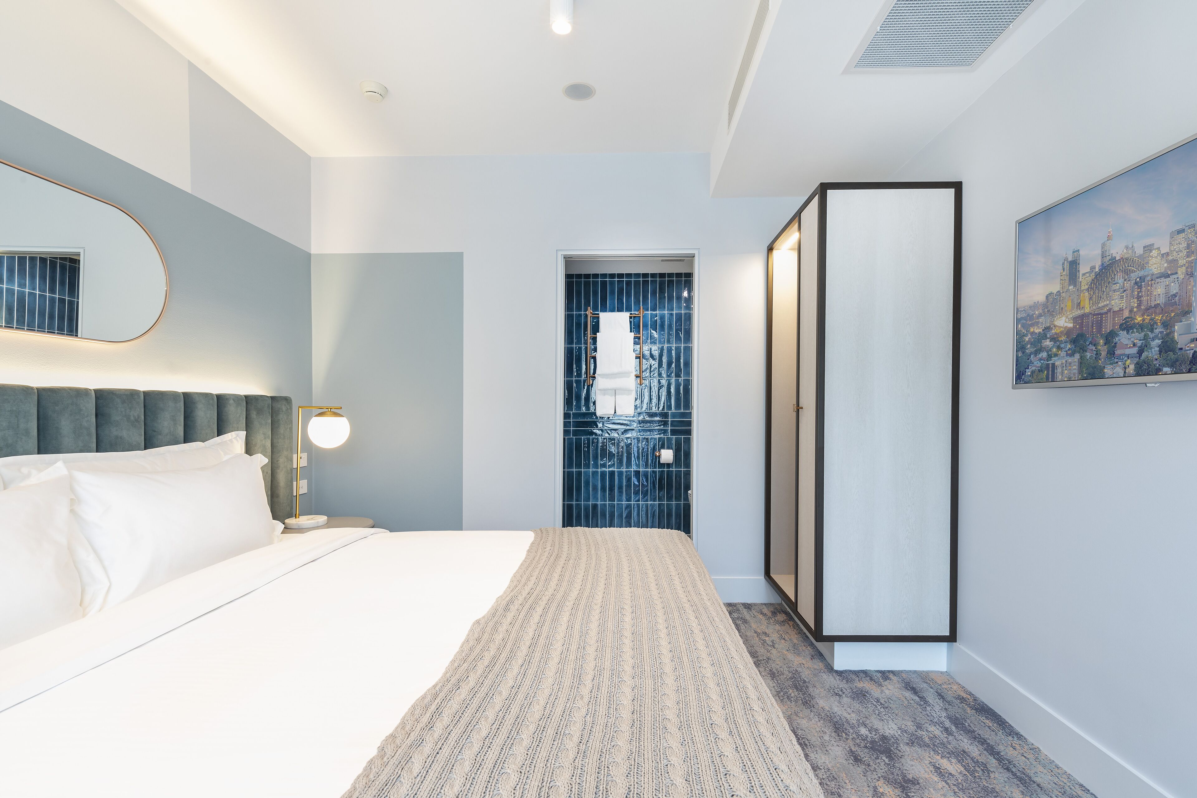 Una habitación de hotel moderna con una cama grande, una mesita de noche, un espejo montado en la pared, un cuadro de paisaje urbano y vistas de la ciudad a través de una ventana.