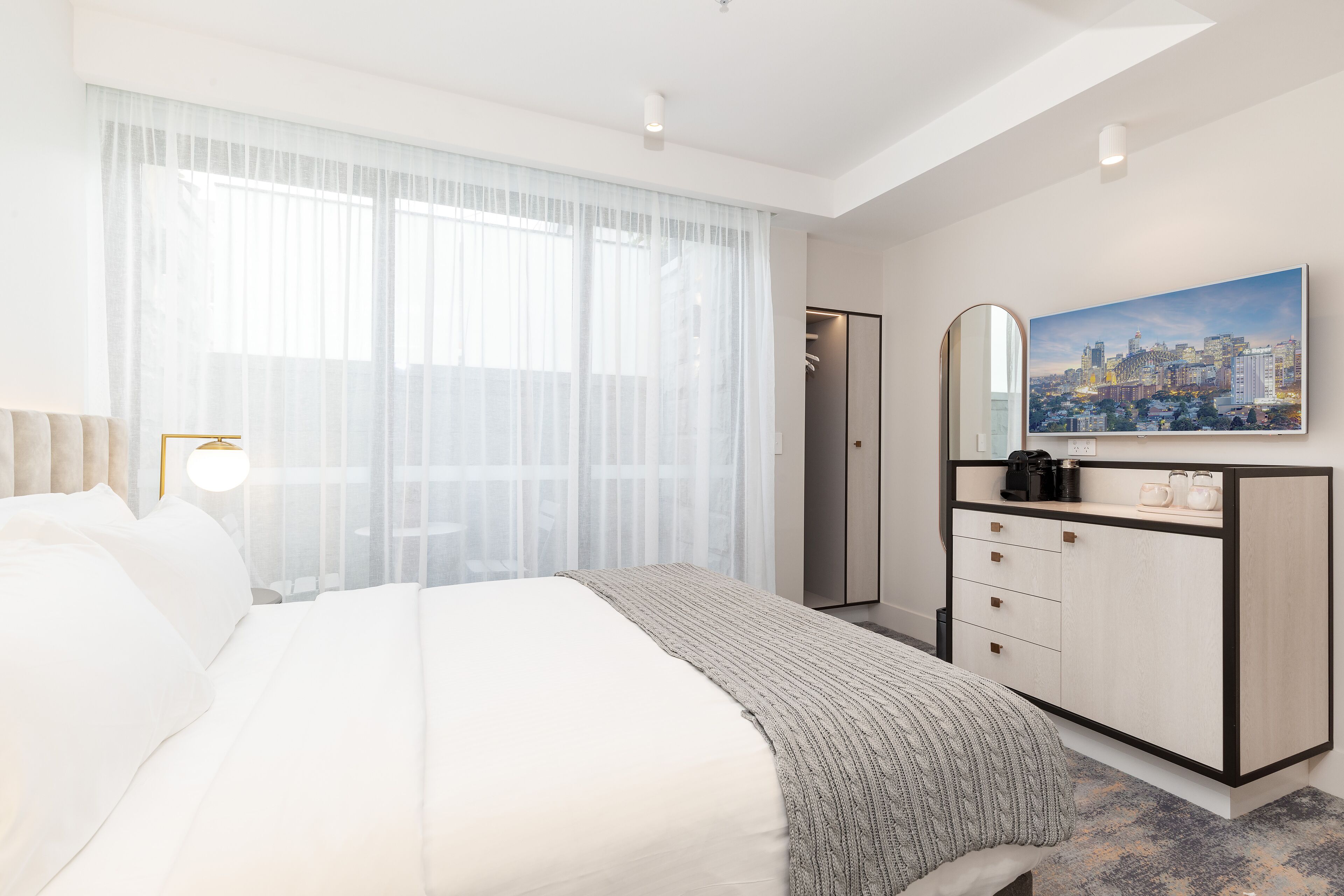 A modern hotel room with a large bed, a wardrobe, a mirror, and a cityscape painting on the wall.