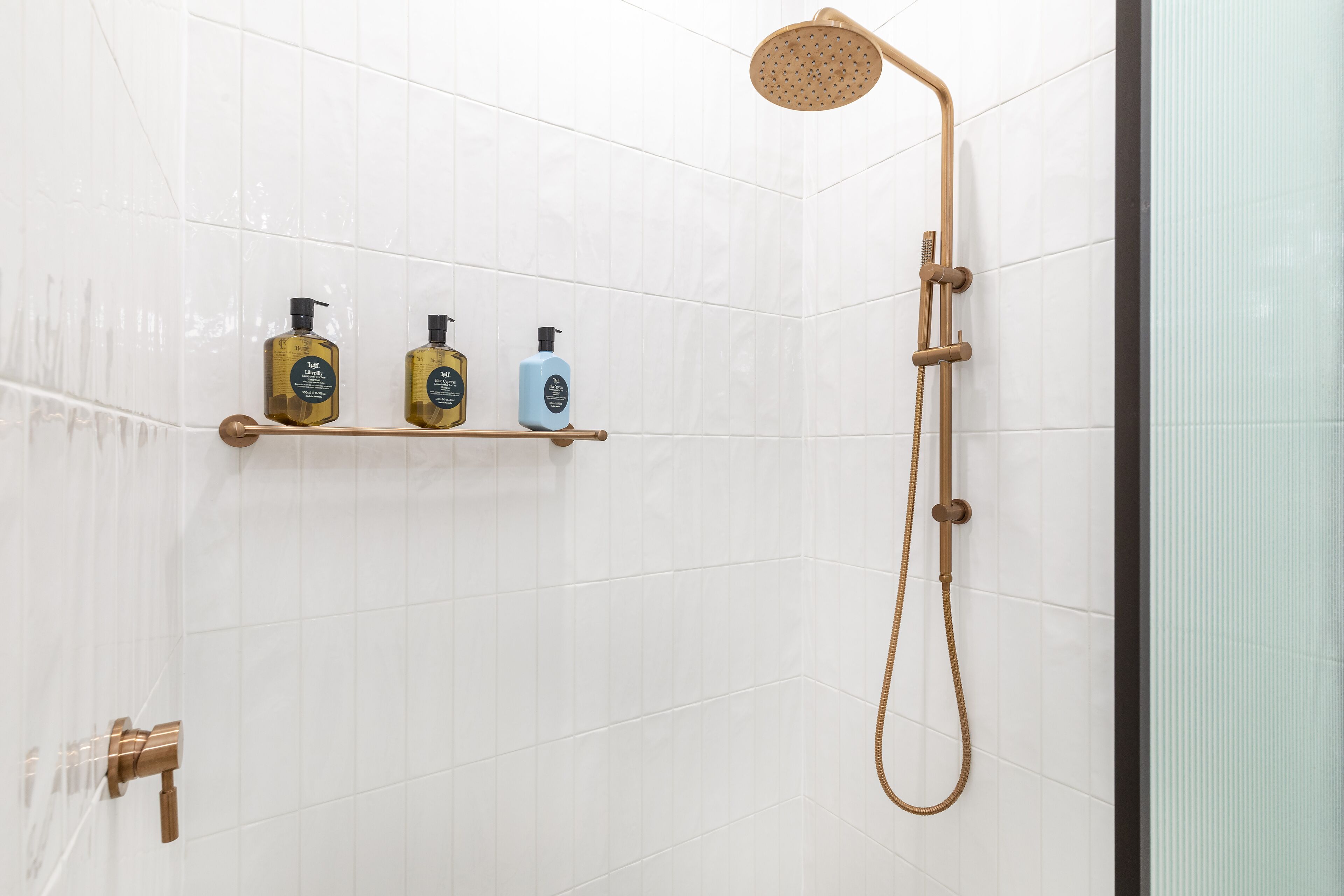 Un espace douche équipé d’un pommeau de douche à main et d’un robinet mural. Trois bouteilles sont posées sur une étagère au-dessus de la douche.