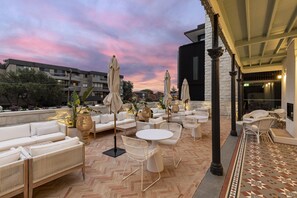 Un patio exterior con mobiliario blanco, que incluye sofás y mesas, bajo una zona techada con un diseño de suelo tipo espiga.