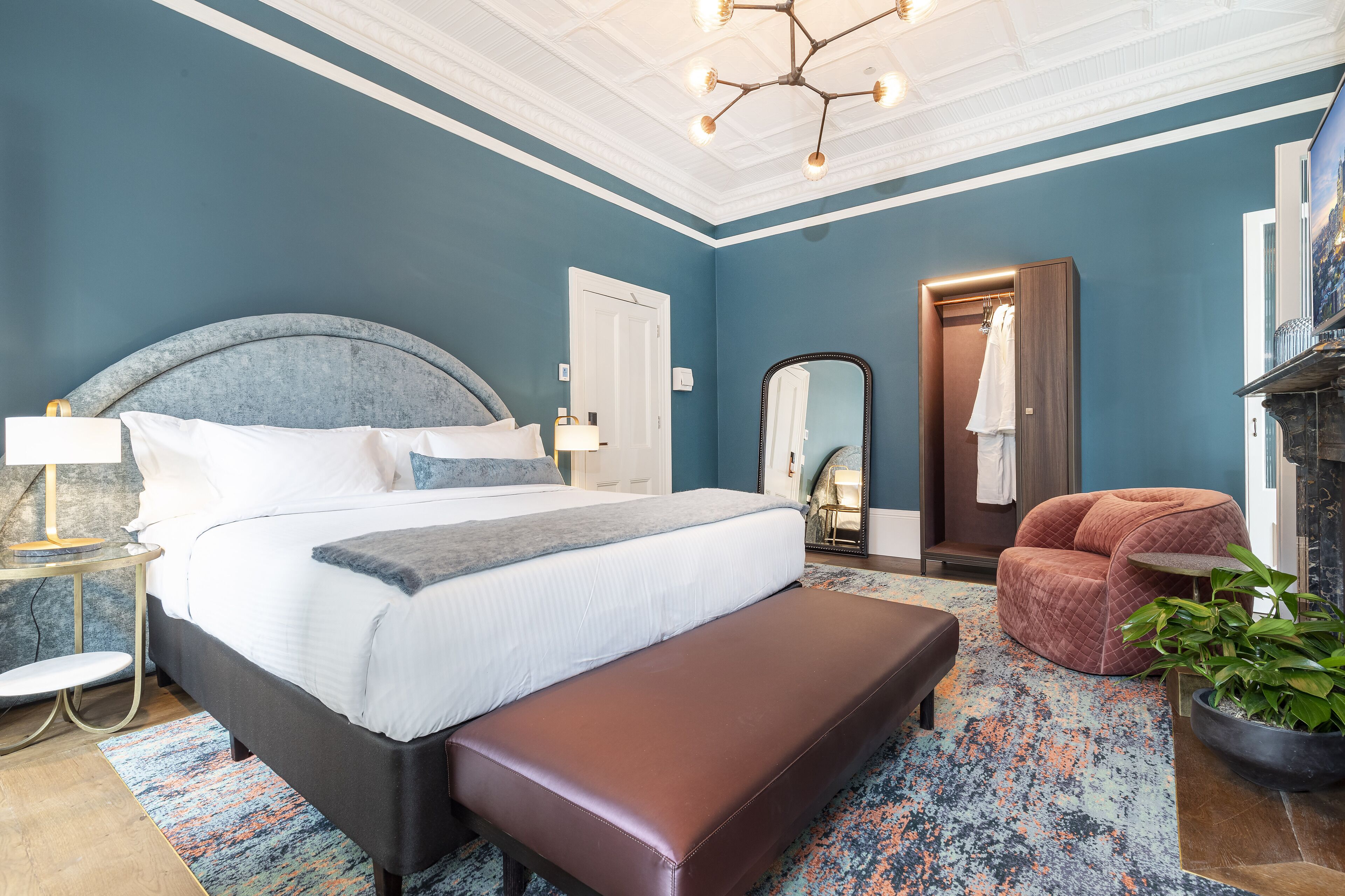 Ein Schlafzimmer mit Bett, Bank, Nachttisch, Lampe, Spiegel, Kleiderschrank und einer Zimmerpflanze.