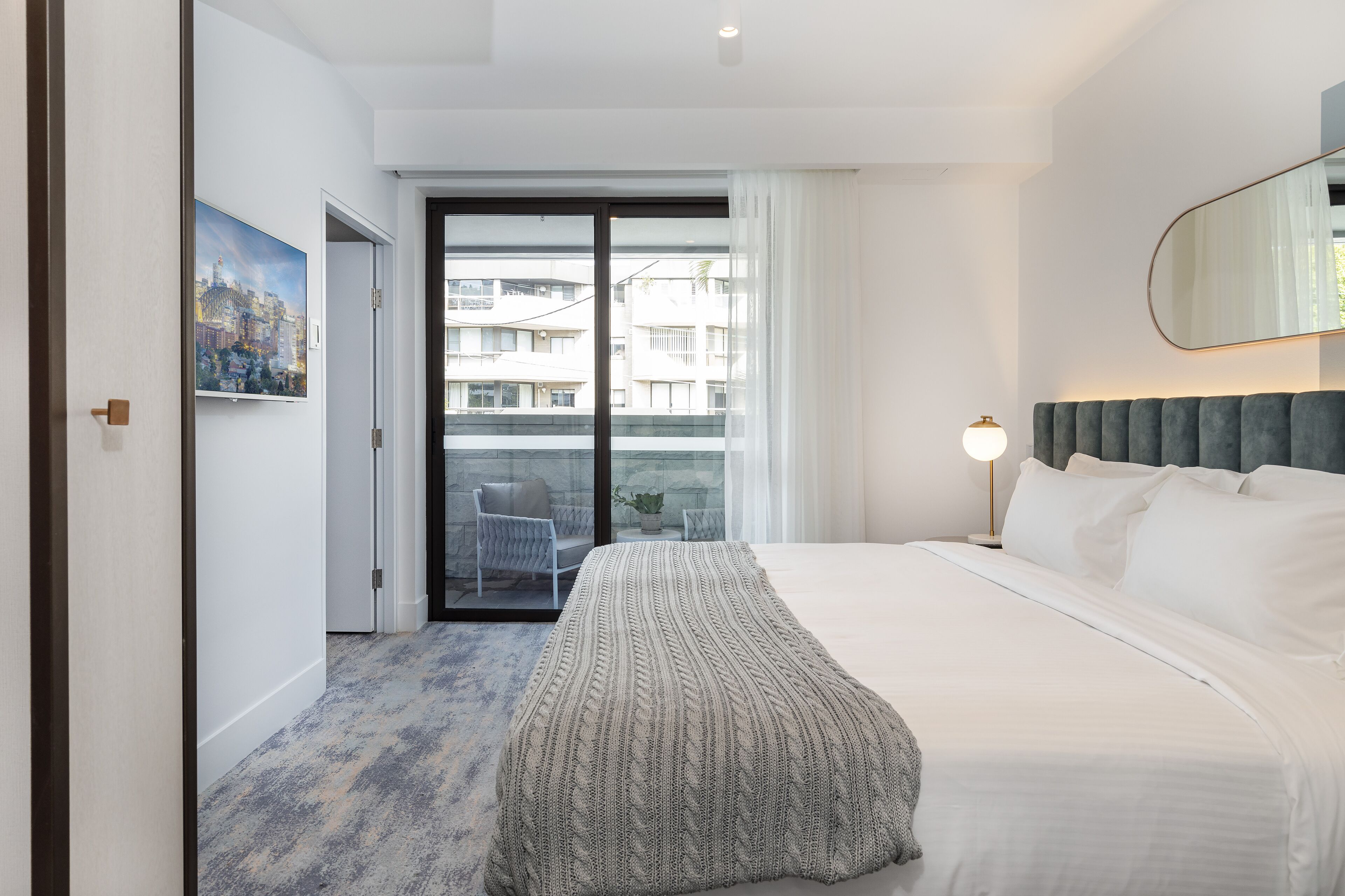 A modern hotel room with a large bed, a view of the city through a sliding door, a painting on the wall, and a bedside lamp.