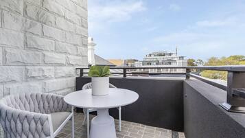 Un balcón con un banco acolchado blanco, una mesita redonda con una planta en maceta y vistas de edificios modernos.