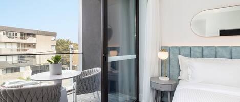 A modern hotel room with a large bed, a glass door leading to a balcony, a small round table with a chair, and a nightstand with a lamp.