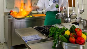 Un chef en una cocina con electrodomésticos de acero inoxidable, diversos vegetales y ollas colgantes.