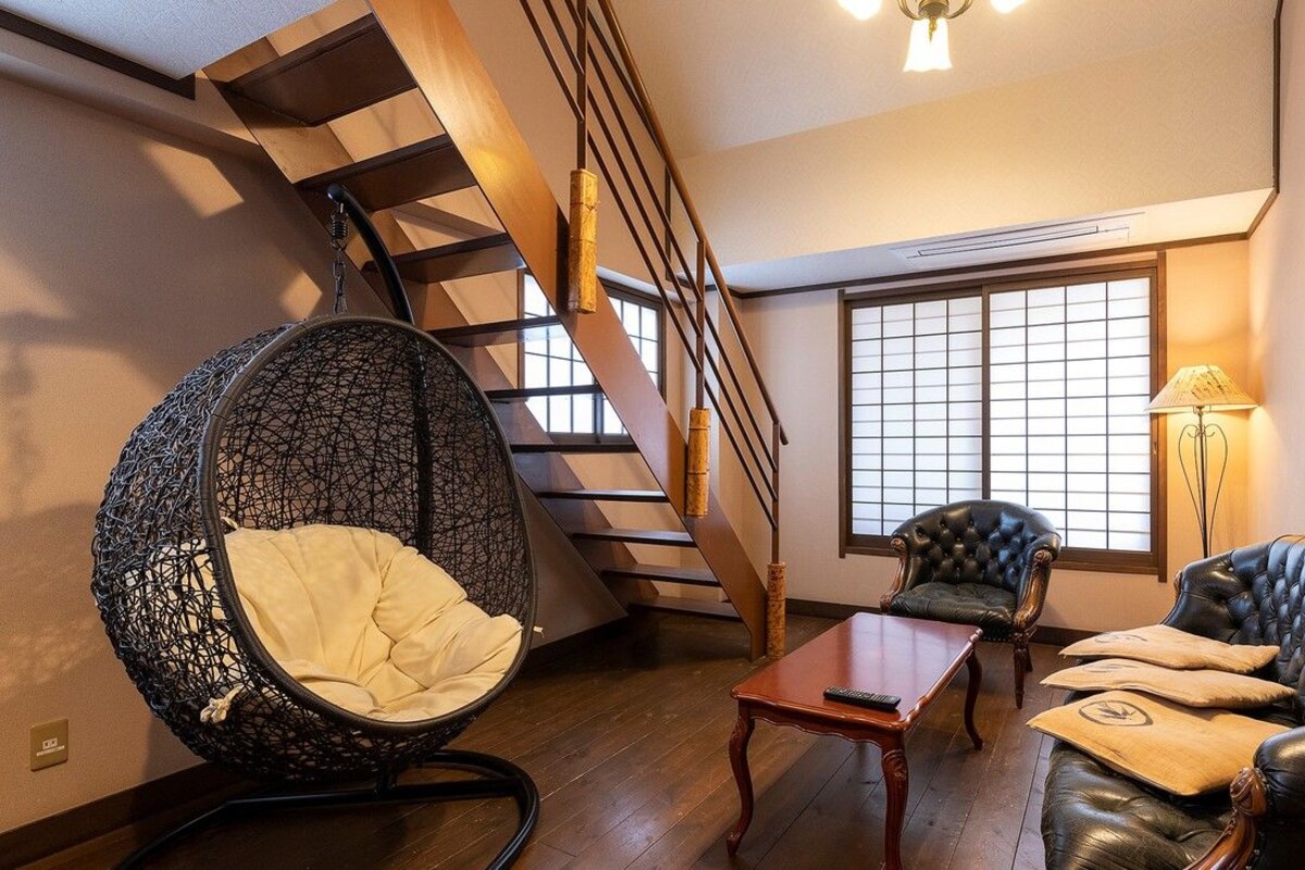 Una habitación con escalera de madera, un sillón mecedor negro, un sofá de cuero, una mesita pequeña y un ventanal con vistas.