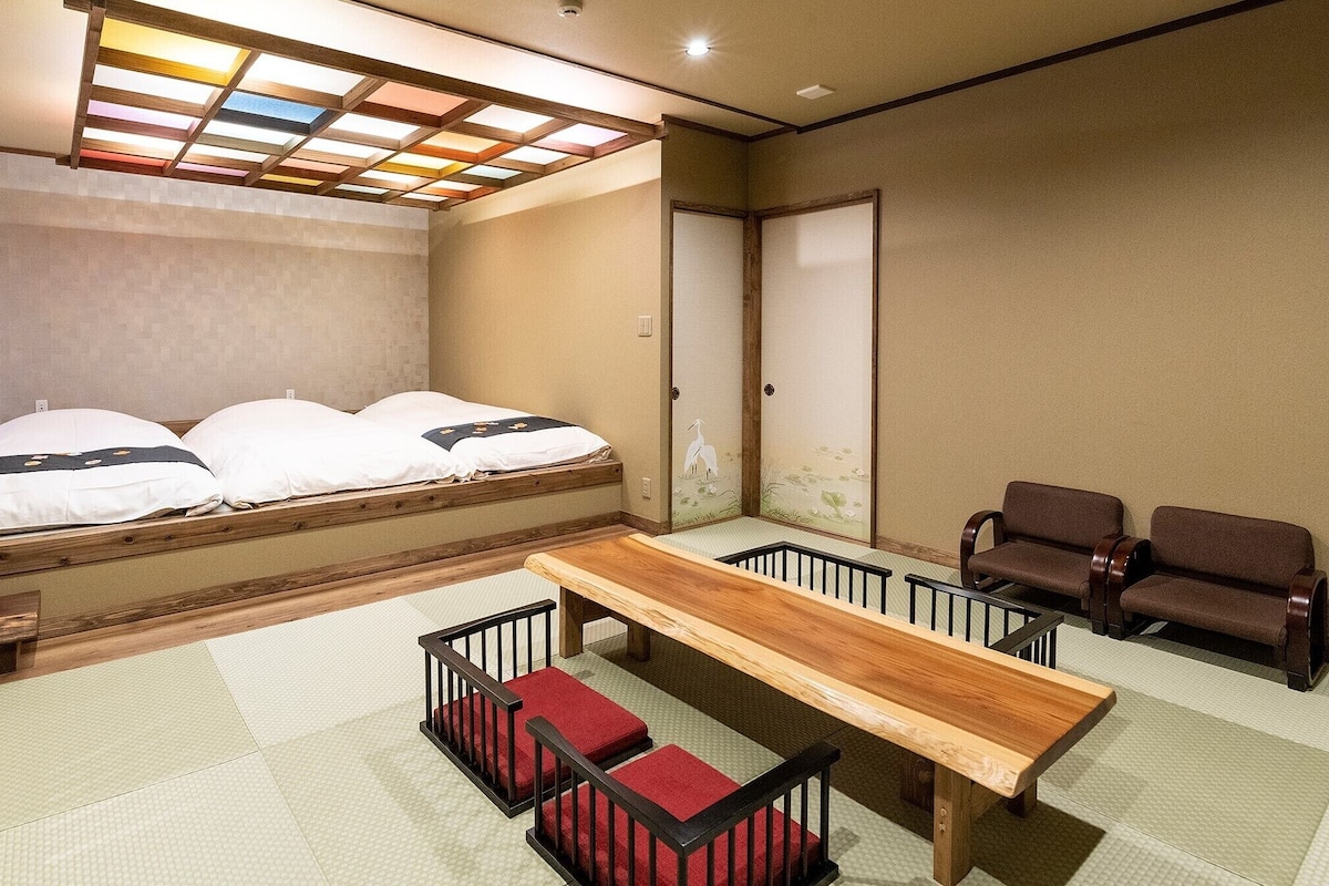 A room with a bed, a wooden table, two chairs, tatami flooring, and a shoji screen door.