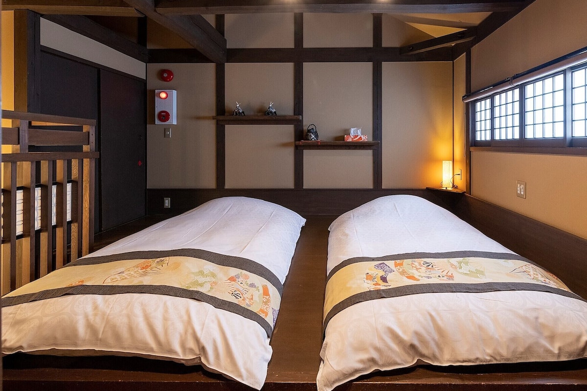 A traditional Japanese-style room with tatami flooring, two single beds with decorative bedding, a sliding door, a small shelf with items, and a window with a grid pattern.