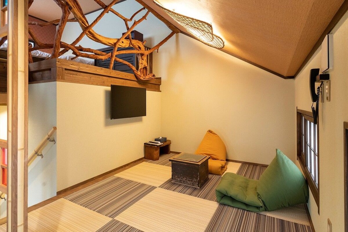 A compact room with a bunk bed, a flat-screen TV, a small table, a green cushion, and a woven mat on the floor.