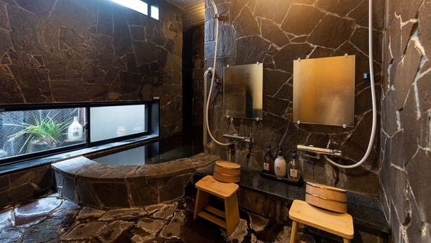 A stone-walled bathroom with a bathtub, a wooden stool, a vanity with bottles, and a window with a view. - Dogo Onsen Atatakai Yado Taniya (Matsuyama)