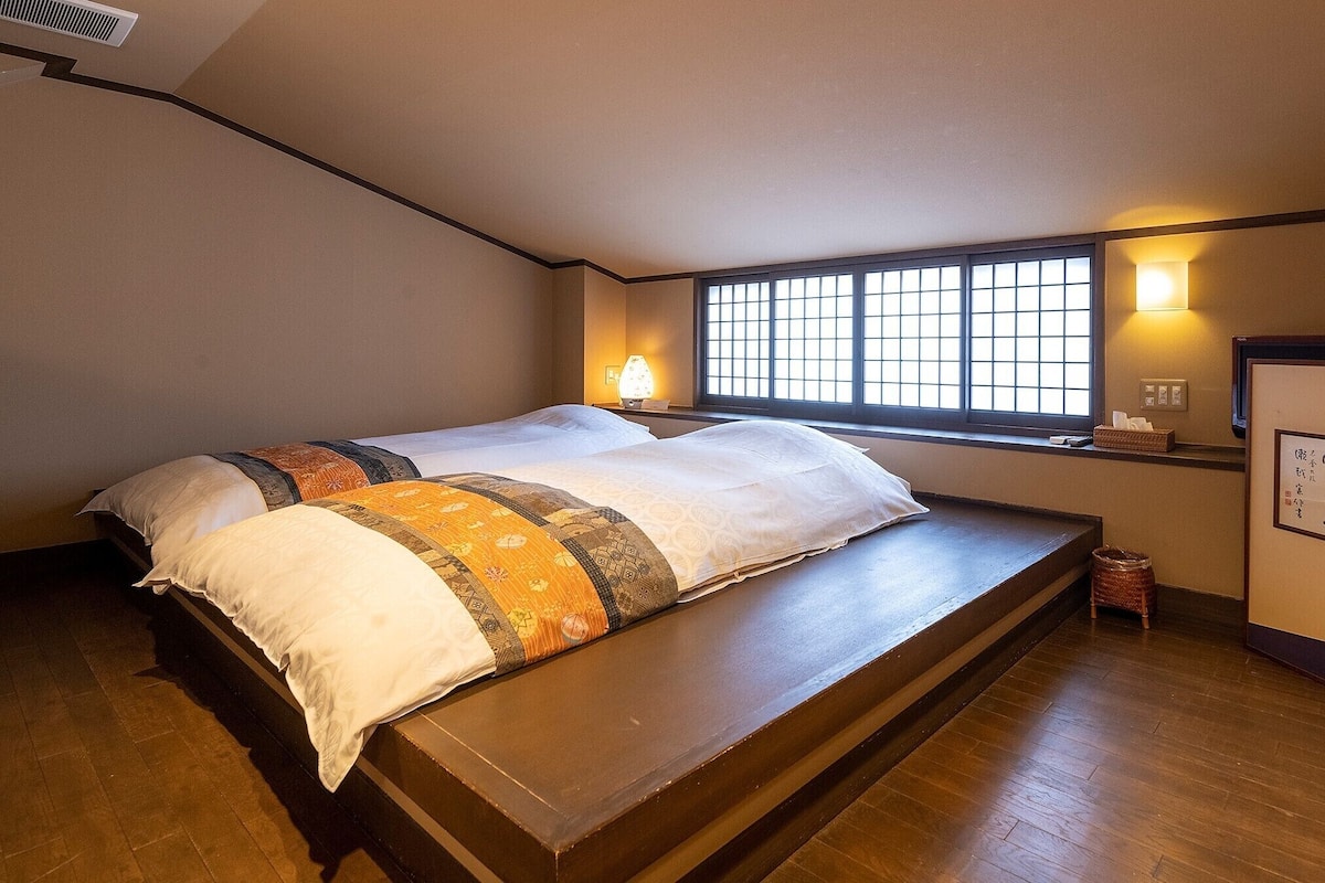 A room with a large bed, a window with a view, a wall-mounted shelf, and a small table.