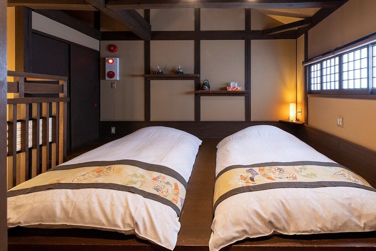 A traditional Japanese-style room with tatami flooring, two single beds with decorative bedding, a sliding door, a small shelf with items, and a window with a grid pattern.