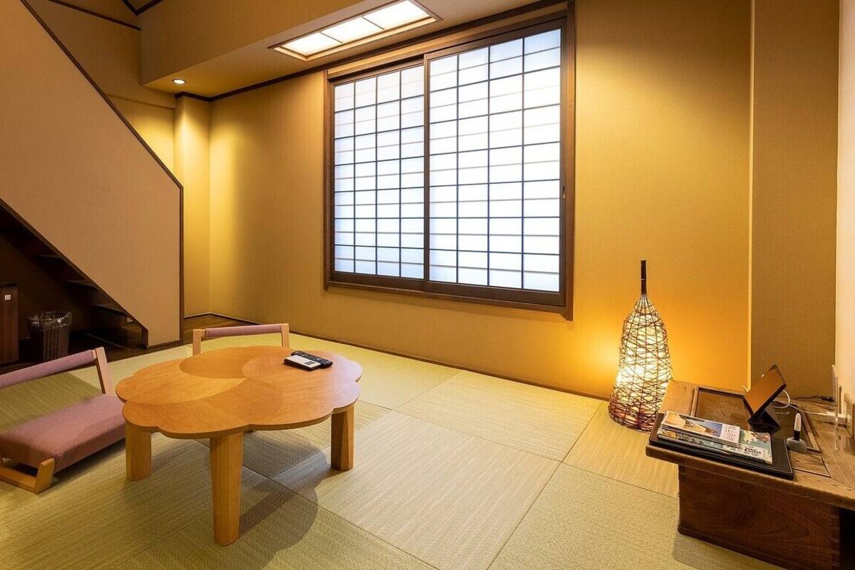 A room with tatami flooring, a low wooden table with chairs, a large shoji screen window, a traditional Japanese paper lantern, and a small wooden cabinet.