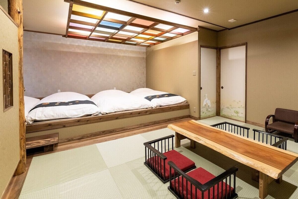 A room with a bed, a wooden table, two chairs, tatami flooring, and a sliding door.