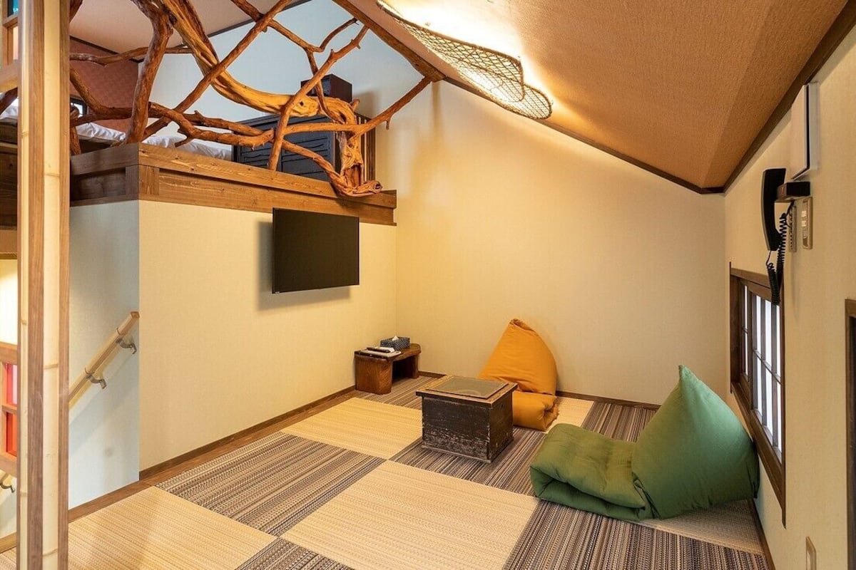 A compact room with a bunk bed, a flat-screen TV, a small table, a green cushion, and a woven mat on the floor.
