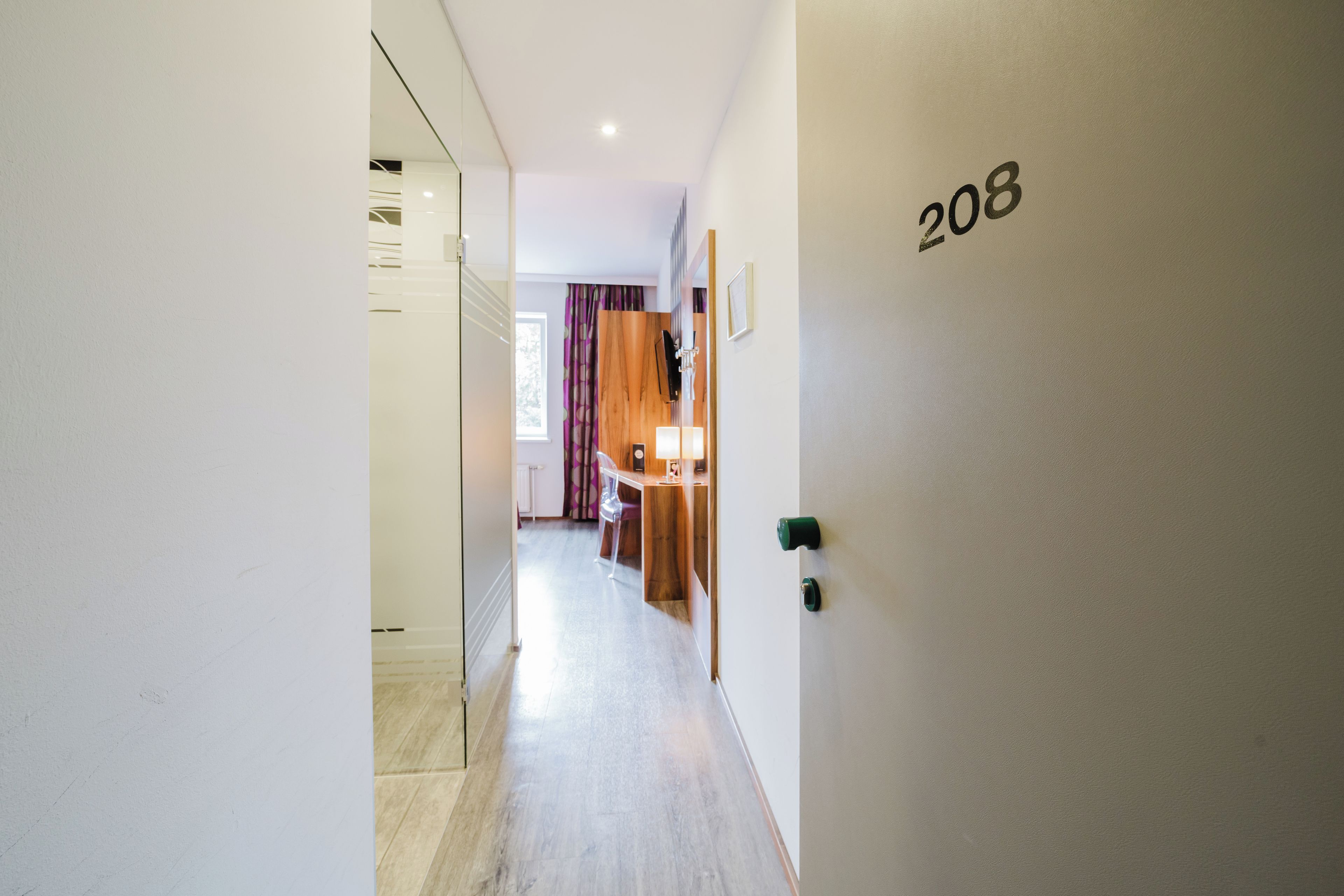 Ein Flur mit einer Tür, auf der '208' steht, die zu einem Zimmer mit Schreibtisch und Stuhl führt.