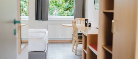 A hotel room with a bed, a desk, and a chair. There is a window with curtains and a view of greenery outside.