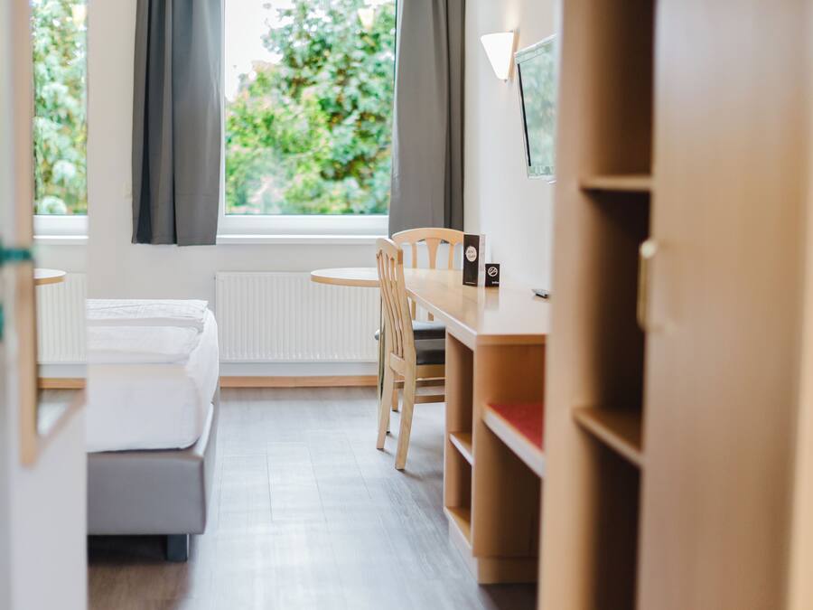 A hotel room with a bed, a desk, and a chair. There is a window with curtains and a view of greenery outside.