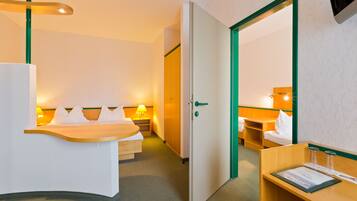 Ein Hotelzimmer mit Bett, Nachttisch und Schreibtisch mit Stuhl. Eine Tür führt in einen weiteren Raum.