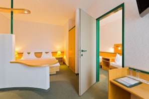 A hotel room with a bed, bedside table, and a desk with a chair. There is a door leading to another room.