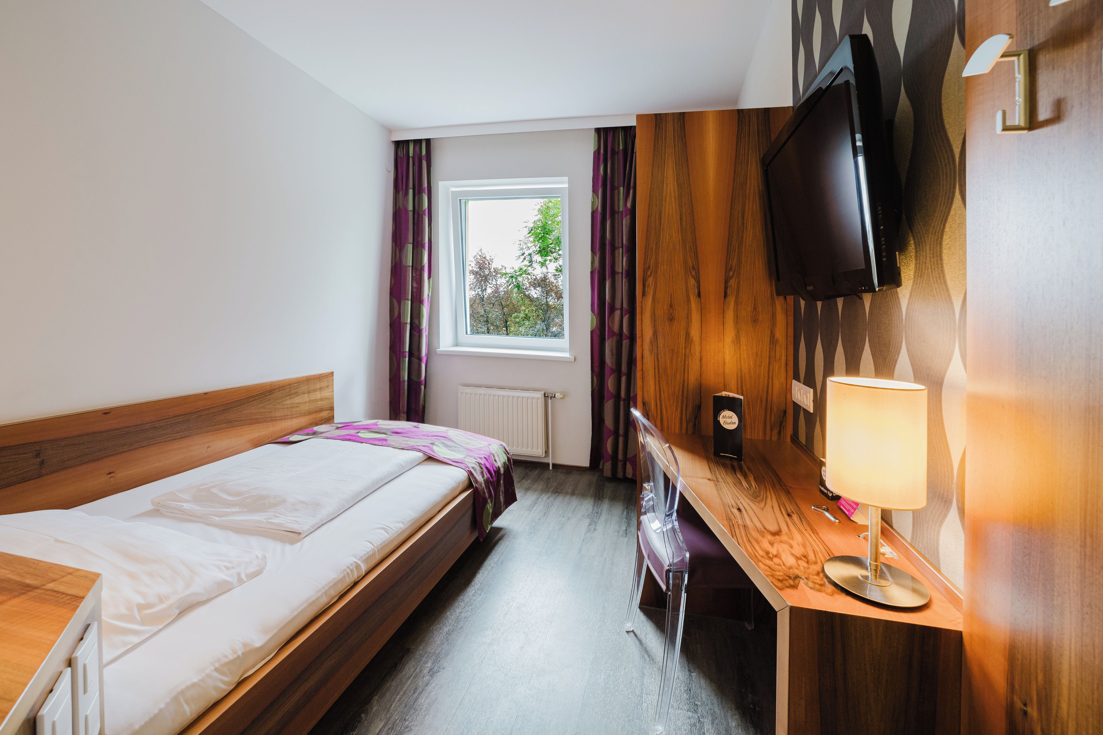 A hotel room with a bed, a desk, a chair, a TV, and a window with curtains.