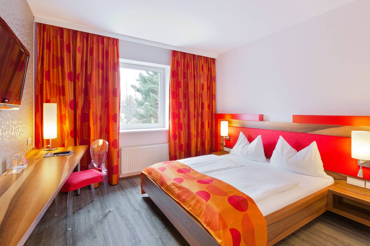 Ein Hotelzimmer mit einem großen Bett, einem Schreibtisch und einem Stuhl. Es gibt ein Fenster mit Vorhängen, einen Nachttisch mit Lampe und einen an der Wand befestigten Fernseher.