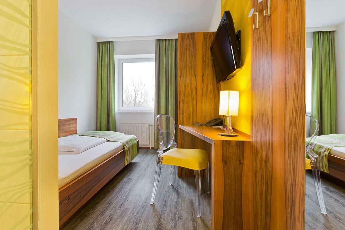 Ein Hotelzimmer mit zwei Betten, einem Schreibtisch mit Stuhl, einem an der Wand befestigten Fernseher und einem Fenster mit Vorhängen.