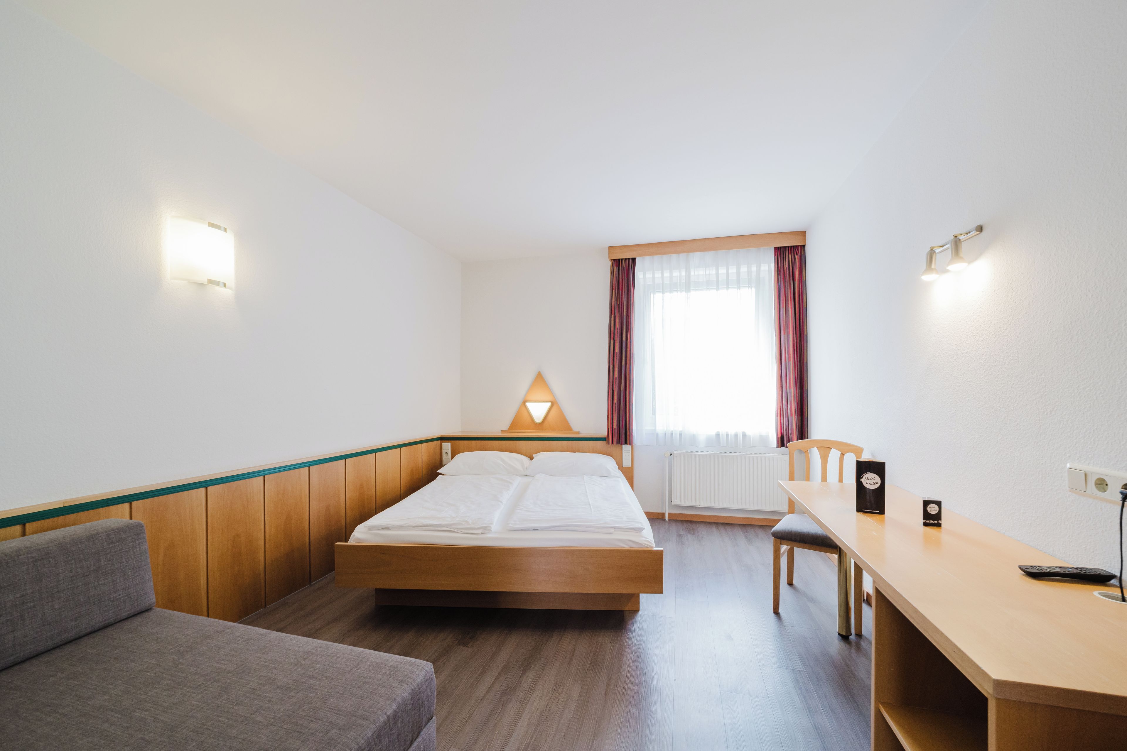 Habitació d'hotel amb llit, escriptori, cadira, làmpada i finestra amb cortines.