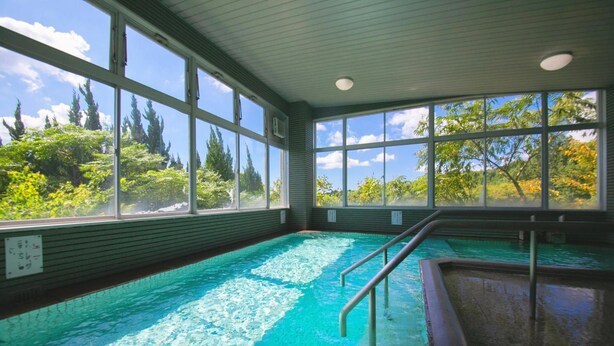 An indoor swimming pool with clear blue water, surrounded by large windows offering views of trees and sky. - Omuta Heights (Omuta)