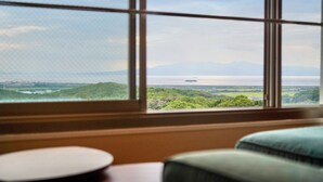 A room with a large window offering a view of a coastal landscape, including a body of water and distant mountains. - Omuta Heights (Omuta)