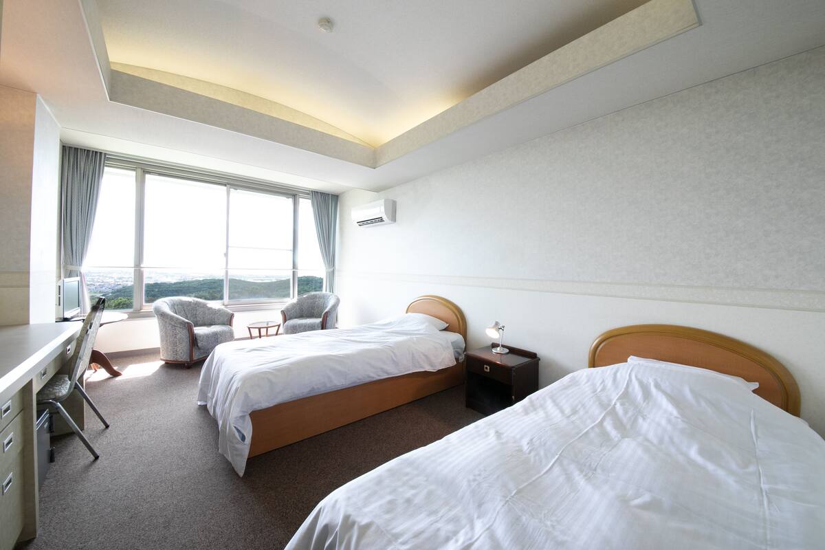 A hotel room with two beds, a desk, a chair, and a large window offering a view.