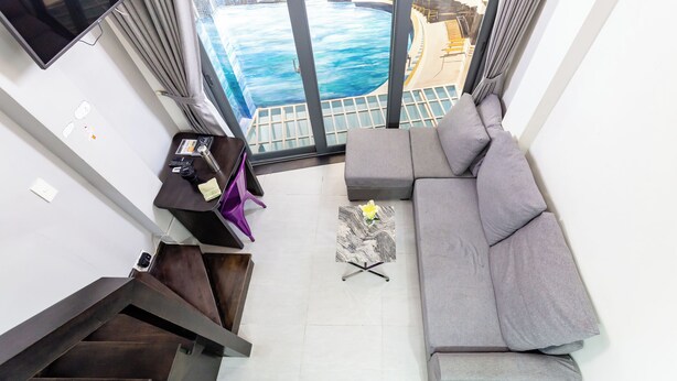 A modern living room with a grey sofa, a small table with a flower, and a view of a pool and buildings through large windows. - The Haven - Lofts by the Park (Ho Chi Minh City)