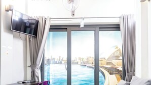 A modern living room with a large sofa, a small table, a purple chair, a TV, and a view of a cityscape through a large window. - The Haven - Lofts by the Park (Ho Chi Minh City)