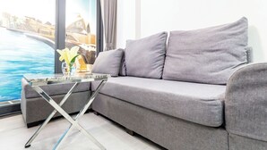 A modern living room with a grey sofa, a glass coffee table, and a view of a body of water through large windows. - The Haven - Lofts by the Park (Ho Chi Minh City)