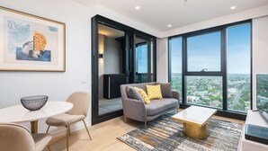A modern living room with a grey sofa, a white coffee table, and a large window offering a city view. - Sky One Apartments by CLLIX (Box Hill)
