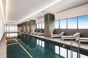 A modern indoor swimming pool with lounge chairs and a view of the city skyline. - Sky One Apartments by CLLIX (Box Hill)